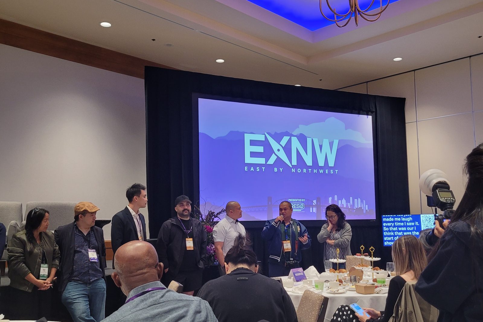 The nine competing teams of the EXNW AI Filmmaking Hack-a-Thon standing together in front of the EXNW East by Northwest screen at the Westin Bayshore in Vancouver
